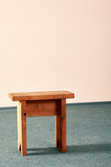 Small wooden stool standing on a gray-green carpet on the floor. Beige empty wall background, copy space. Nobody.