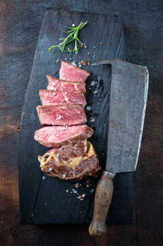 Barbecue dry aged angus rib eye beef steak sliced offered as top view with a rustic old kitchen cleaver on an old charred wooden board