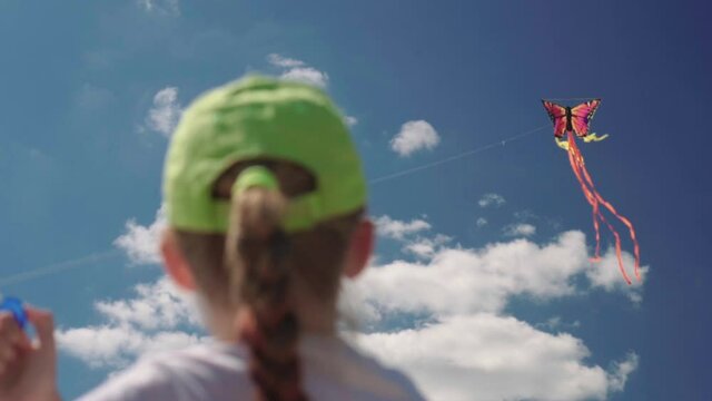 Pretty child girl in green cap playing with kite on clear summer day. Blue sky and clouds. Summer holiday and childhood concept. To be in control of a colourful kite flying in the wind slow motion