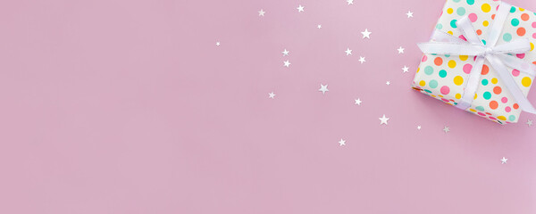 Gift box decorated with white bow and paper with colored polka dots on pastel background with star confetti. Selective focus, festive banner