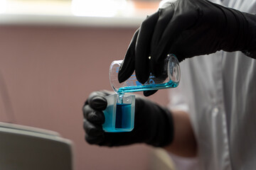 Chemical experiment in a laboratory: hands in rubber gloves are pouring blue liquid to a glass flask, close up