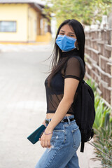 Fototapeta premium beautiful young latin woman with long black hair, sideways and with a cell phone in her hand, wearing a mask, transparent blouse with jeans and a black backpack, on a path outside