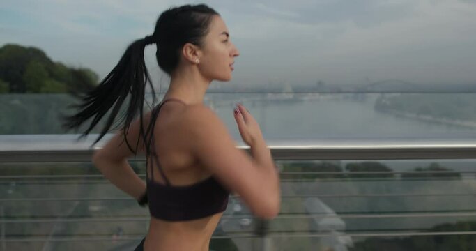 Strong sportswoman running fast on bridge. Tracking shot of fit female in sportswear running on modern bridge during fitness training in evening in city