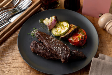 Roasted beef fillet, dark and yummy, with grilled vegetables as a side dish, knife, fork and pepper shaker aside on a wooden table
