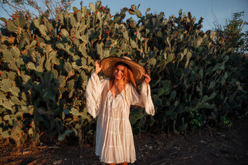 Young woman dressed in white dress and straw hat  standing among cactuses, smile and look at the camera