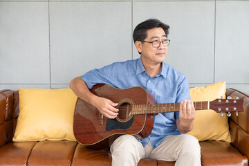 Asian elder man playing guitar at the sofa inside of the house. Active senior lifestyle after...
