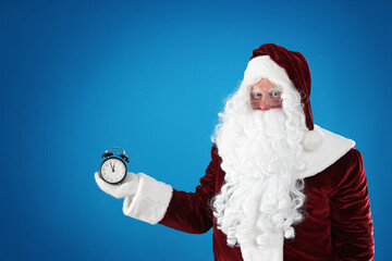 Fototapeta na wymiar Santa Claus holding alarm clock on blue background, space for text. Christmas countdown