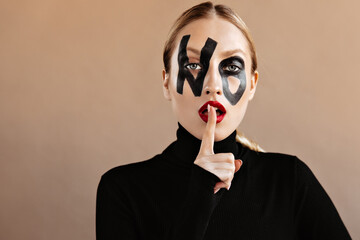 Gray-eyed woman with painted word no on her face puts index finger to her red lips on isolated background
