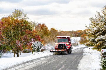 View of red truck with snowplow plowing suburban Midwestern street after first snowfall; some trees preserve their colorful fall leaves; Missouri