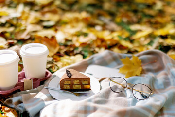 picnic in the autumn park. A piece of chocolate cake with coffee to go on a blanket next to glasses and a smartphone against a background of maple leaves.