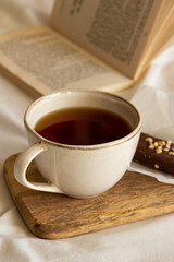 Cup with tea and a chocolate bar on a wooden board next to an open book on a white bed
