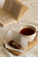 Cup with tea and a chocolate bar on a wooden board next to an open book on a white bed