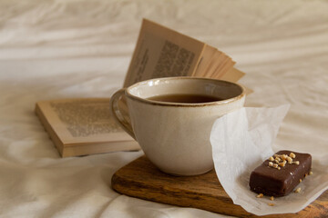 Cup with tea and a chocolate bar on a wooden board next to an open book on a white bed
