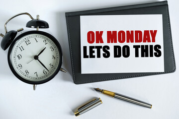 On the table there is a clock, a pen, a notebook and a card on which the text is written - OK MONDAY LETS DO THIS