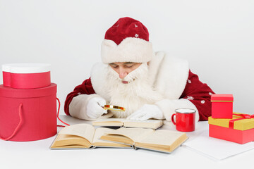 A tired Santa Claus sits at a table and studies books using a magnifying glass. There are Christmas gifts on the table.