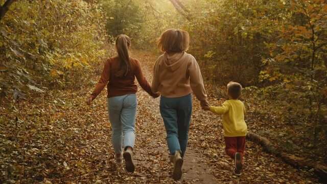 Back view of a young mother holding children by the hands happily running through the autumn forest