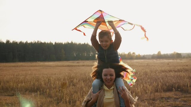 Happy little boy holding a kite in hands while sitting on the shoulders of mom cheerfully run on the meadow, slow motion