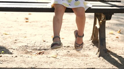 Little child girl waggle sway legs as she sit on a bench in a park enjoy in good mood because of eating ice cream