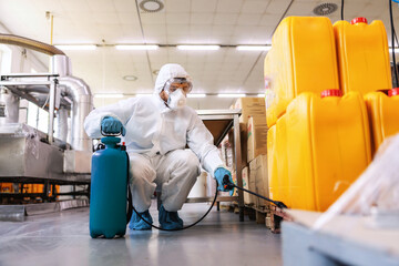 Worker in sterile uniform with rubber gloves holding sprayer with disinfectant and spraying around warehouse. Corona outbreak concept.