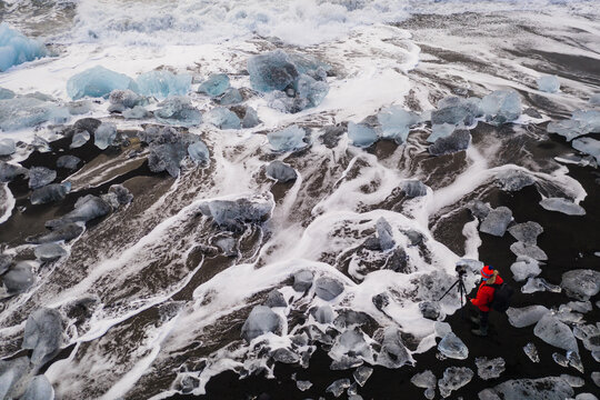 Red clothed man who photographing waves between ice rocks in famous Icelandic black beach. Black beach, witch ice diamonds landscape to your calendar or postcard.