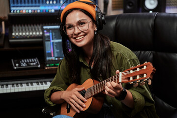 Fototapeta na wymiar Portrait of beautiful young brunette, female artist smiling aside, playing ukulele while sitting in recording studio