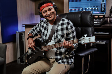 Obraz premium Portrait of happy young man, male artist in headphones smiling aside, playing guitar while sitting in recording studio