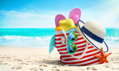 Fototapeta na wymiar Summer beach bag and accessories - straw hat, flip flops and sunglasses on sandy beach and azure sea on background
