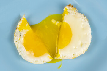 An overhead view of a fried egg cut in half, flowing yolk onto a blue plate. Wood background.