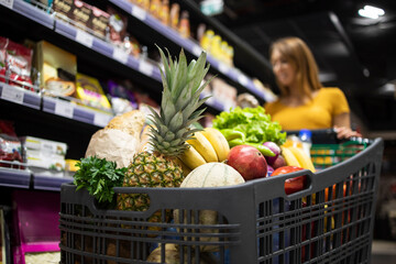 Supermarket shopping. Close up view of shopping cart overloaded with food while in background female person choosing products. © littlewolf1989