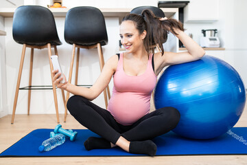 Young pregnant female using mobile device during working out in living room at home. Caucasian woman sitting on blue exercise mat doing photo by phone. Bottle of water, dumbbells on the mat