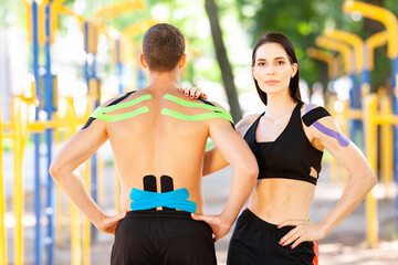 Portrait of brunette woman holding hand on shoulder of unrecognizable man, professional caucasian athletes with kinesiological taping on bodies, posing at sports ground, looking at camera.