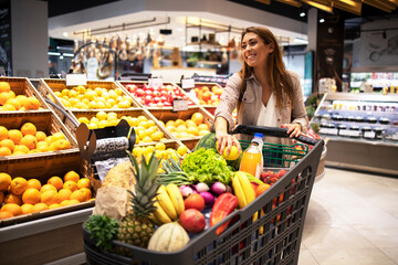 Beautiful smiling woman pushing shopping cart and taking fruits off the shelves in supermarket. Buying food at grocery store. - obrazy, fototapety, plakaty