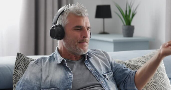 happy excited caucasian adult man resting on couch wear wireless headphones listen to music playing imaginary guitar,mature latino male sitting on sofa having fun playing his favorite song