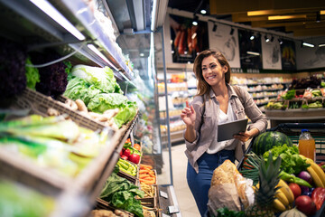 Brunette woman enjoys shopping food at supermarket. Choosing best vegetables on the shelf in grocery store. - obrazy, fototapety, plakaty