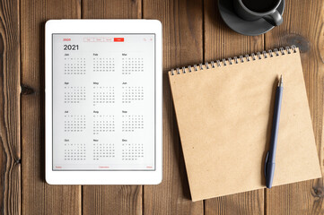 a tablet with an open calendar for 2021 year, a cup of coffee and a craft paper notebook on a wooden boards table background