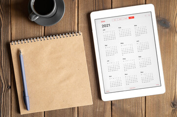 a tablet with an open calendar for 2021 year, a cup of coffee and a craft paper notebook on a...