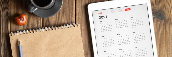a tablet with an open calendar for 2021 year, a cup of coffee, chestnuts and a craft paper notebook on a wooden boards table background. banner