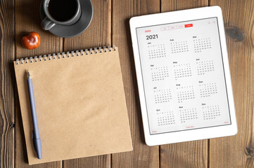 a tablet with an open calendar for 2021 year, a cup of coffee, chestnuts and a craft paper notebook on a wooden boards table background