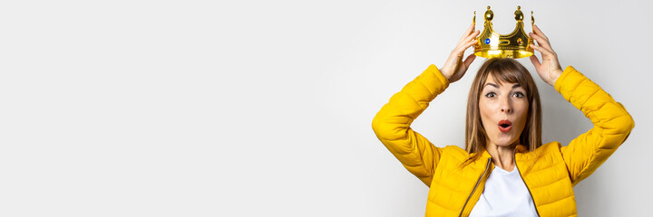 Young woman in a yellow jacket puts a golden crown on her head on a light background. Emotion laughter, surprise, kiss. Concept of a queen, luck, the best, win, victory. Banner © Alex