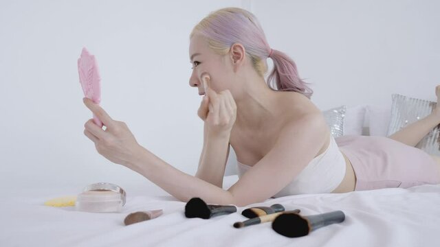 Beauty blocker pretty Asian woman make up by luxury and softy blush for take care skin health with small pink mirror. Modern blonde and pink hair female model on the bed wear sexy white bra