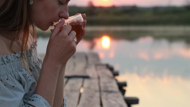 A Young Girl Drinking Hot Tea at the Sunrise on the Wooden Pier in Nature Lake. Outdoor in Sunlight, Enjoying her Morning Tea and enjoys the scenery in the lake