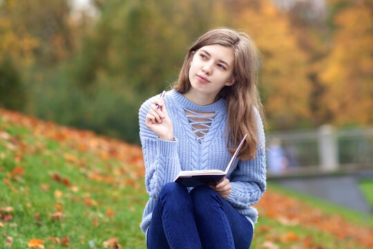 Young beautiful happy woman, teenager student is studying outdoors in park writing in notebook or diary, with thoughtful pensive look, doing homework, dreaming. 