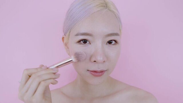 Close up young face Asian woman make up by luxury and softy blush for take care skin health. Modern blonde and pink hair beautiful female model on isolate pink background wear sexy white bra