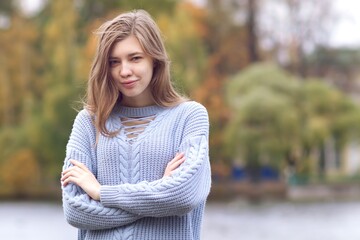 Obraz premium Portrait of young beautiful woman standing in autumn park in sweater with her hands crossed, smiling, looking at camera 