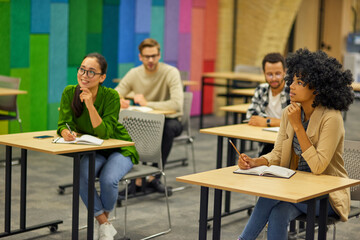 Group of young multiracial people sitting at desks in the modern office and listening to coach or speaker during corporate team building training or business seminar