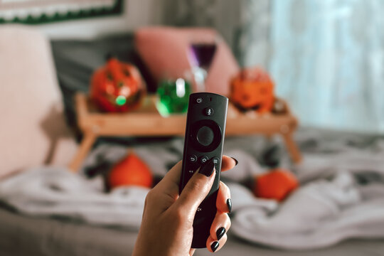 Woman Hand With Black Manicure Holding Tv Remote With Halloween Decor In Background, Halloween Movie Night Concept