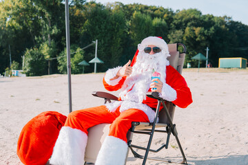 Santa Claus in sunglasses drinks a cocktail while lying on a sun lounger on the lake beach. Santa in the tropics