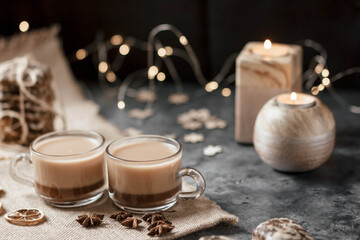 Two identical cups of hot cocoa are on the table. Under them is a napkin made of natural material, next to it lies star anise and slices of dried lemon.