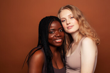 two pretty girls african and caucasian blond posing cheerful together on brown background,...