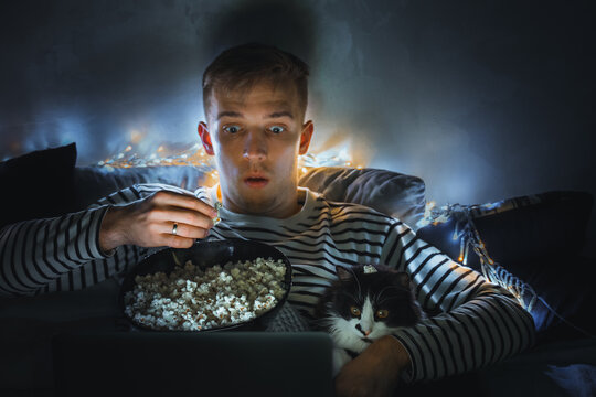 Young Man With Black Cat Watching A Movie Eating Popcorn On TV At Home. Movie Night. Relax,rest Watching A Horror Film Or Video On Screen. Background Lighting. Fun Scared Excited People On The Couch.